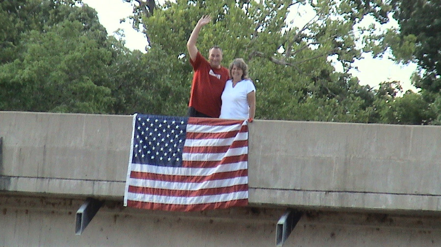 A man in red shirt and woman in white shirt stand behind a concrete barrier. An American flag is draped over the concrete barrier.