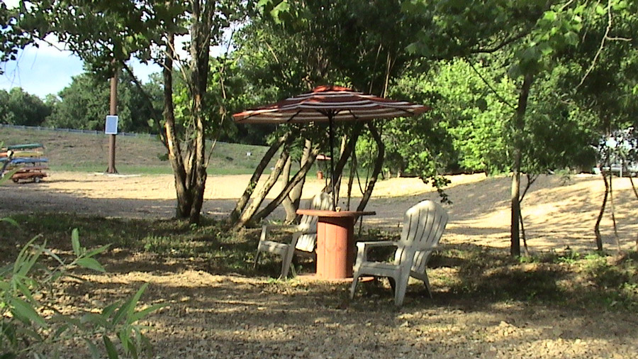 A white chair near a wood spool table and umbrella on gravel under shady trees. 