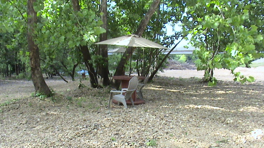 A white chair near a wood spool table and umbrella on gravel under shady trees. 